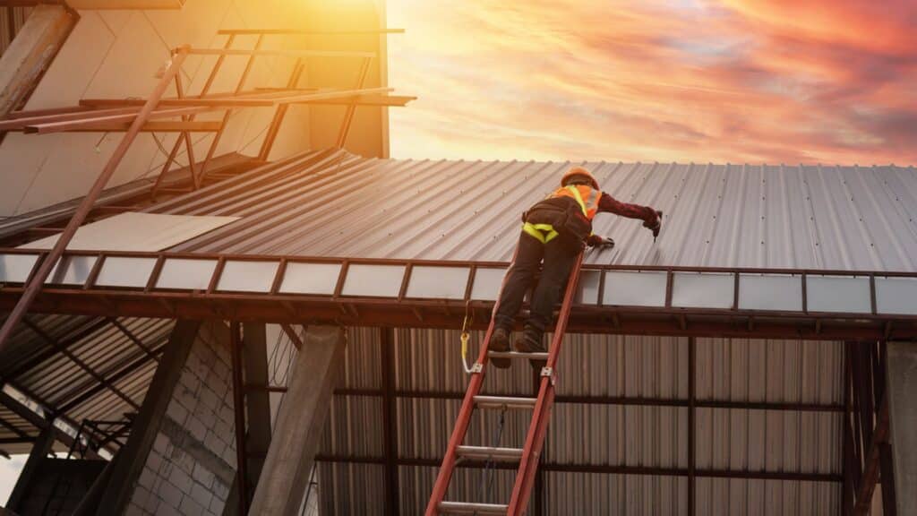 A worker in a safety vest climbs a ladder to work on the metal roof of a building